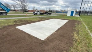 Rectangular freshly poured smooth concrete shed pad set in a graded lawn area with NG Masonry truck on the access road, serving Victor NY