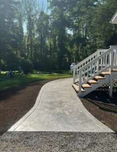 Curving light gray slate textured stamped concrete walkway leading from a gravel driveway to a covered porch with white railings in a wooded yard, by NG Masonry serving Batavia NY