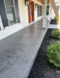 Wide charcoal slate textured stamped concrete porch slab running along a white sided home with dark shutters and a red front door, by NG Masonry serving Batavia NY