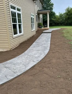 Curving light gray ashlar slate stamped concrete walkway through fresh landscaping leading to a covered porch at a tan sided home, by NG Masonry serving Brighton NY