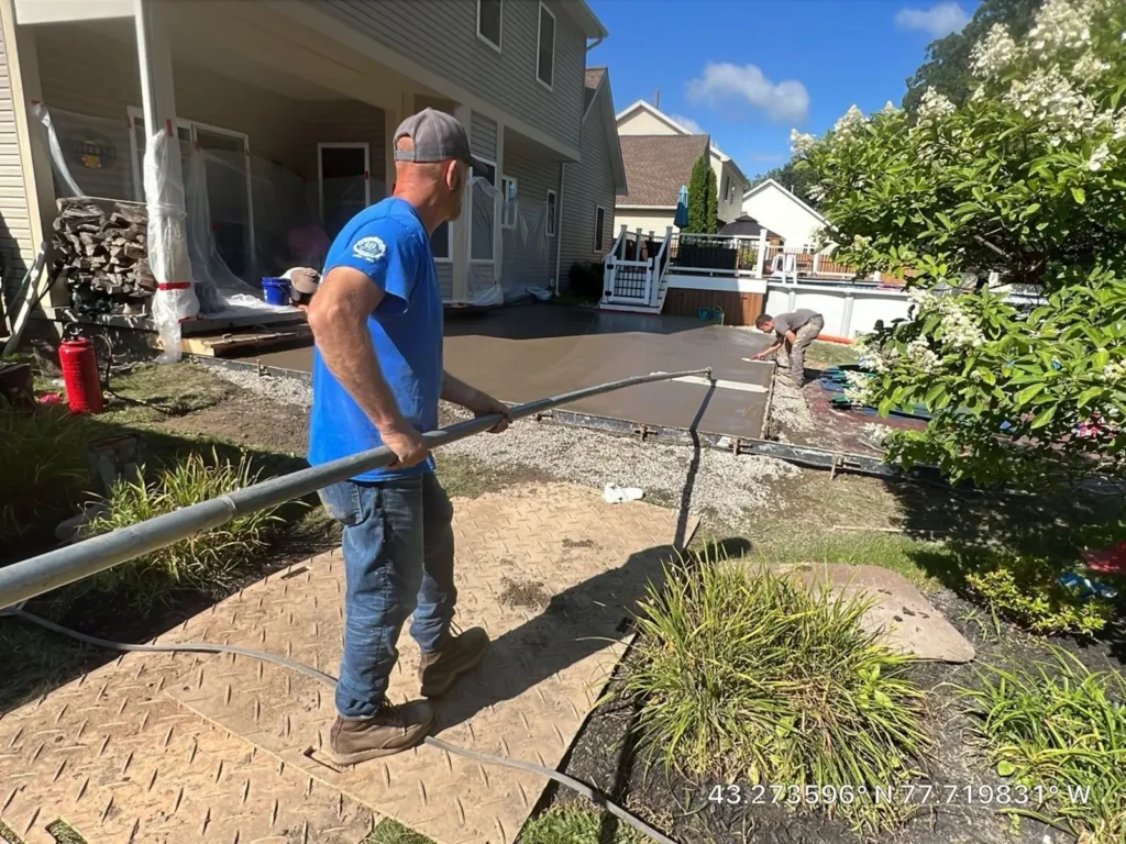 NG Masonry worker using a vibratory screed during a fresh stamped concrete patio pour beside a tan sided home, serving Canandaigua NY