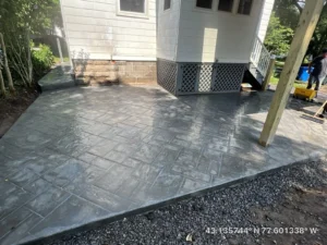 Gray ashlar slate stamped concrete patio under a covered deck with white lattice skirt at a sided home, by NG Masonry serving Farmington NY
