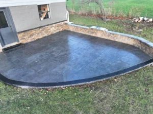 Charcoal slate textured stamped concrete patio with stone seat wall behind a gray sided home, by NG Masonry serving Marion NY