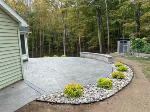 Light gray slate textured stamped concrete patio with stone seat wall in a wooded backyard, by NG Masonry serving Mendon NY
