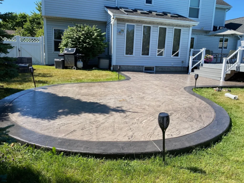 Curved gray slate textured stamped concrete patio with charcoal border at a green sided home, by NG Masonry serving Newark NY