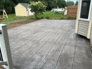 Wide gray wood plank stamped concrete patio in a sunny backyard with playset and shed at a cream sided home, by NG Masonry serving Pittsford NY