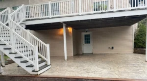 Light gray slate textured stamped concrete patio under a deck with white staircase at a sided home, by NG Masonry serving Rochester NY