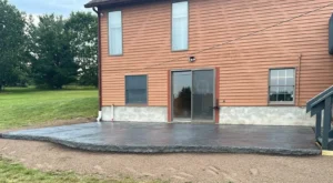 Gray slate textured stamped concrete back patio with rough rock faced edge behind a red sided home, by NG Masonry serving Victor NY