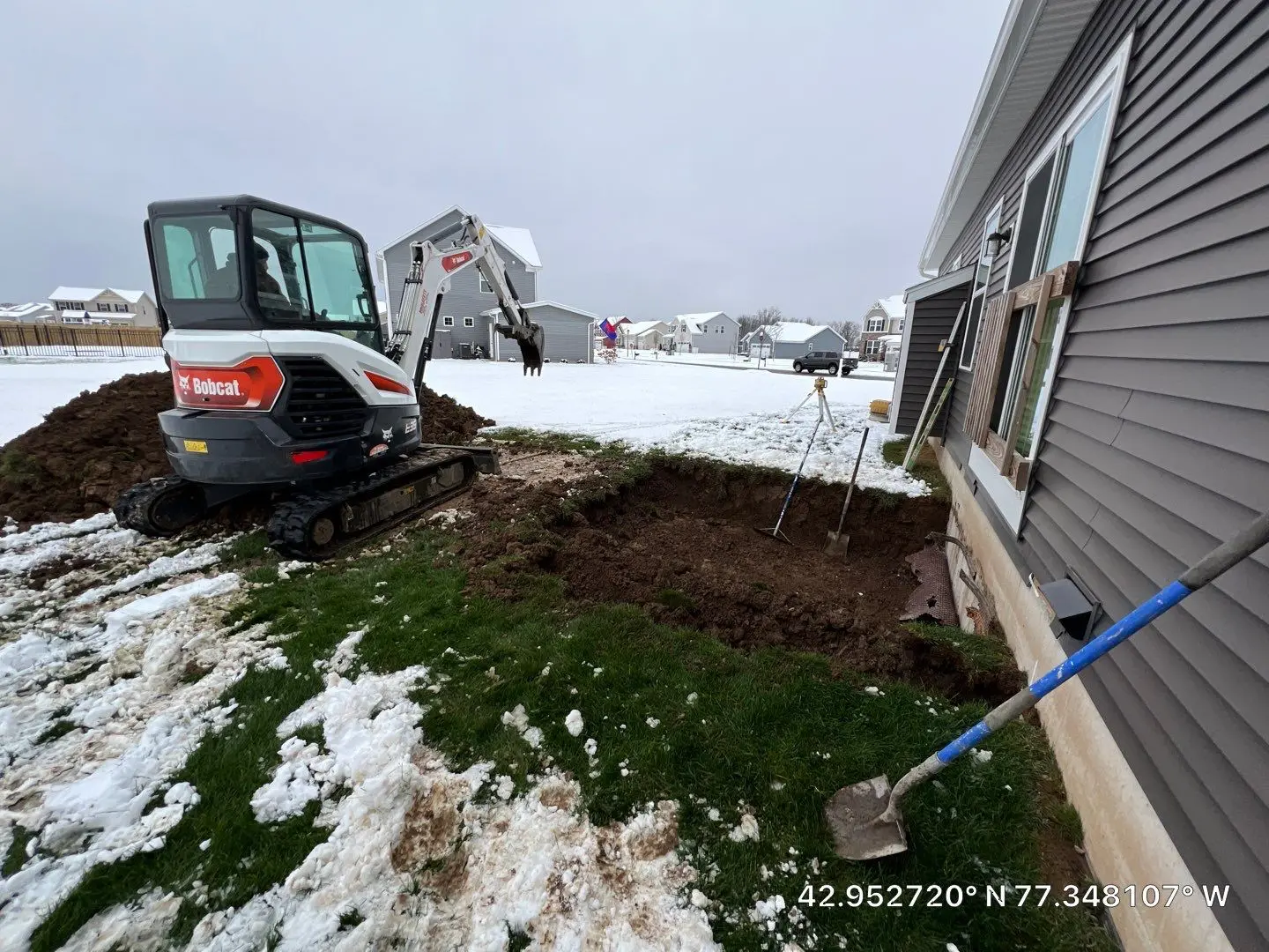 Bobcat mini excavator on a snowy lot beside a gray sided home with a fresh foundation excavation dug along the side wall, by NG Masonry serving Farmington NY