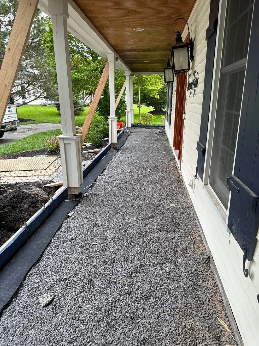 Compacted gravel base inside wood form boards under a wood ceiling covered porch ready for a new concrete porch pour, by NG Masonry serving Rochester NY