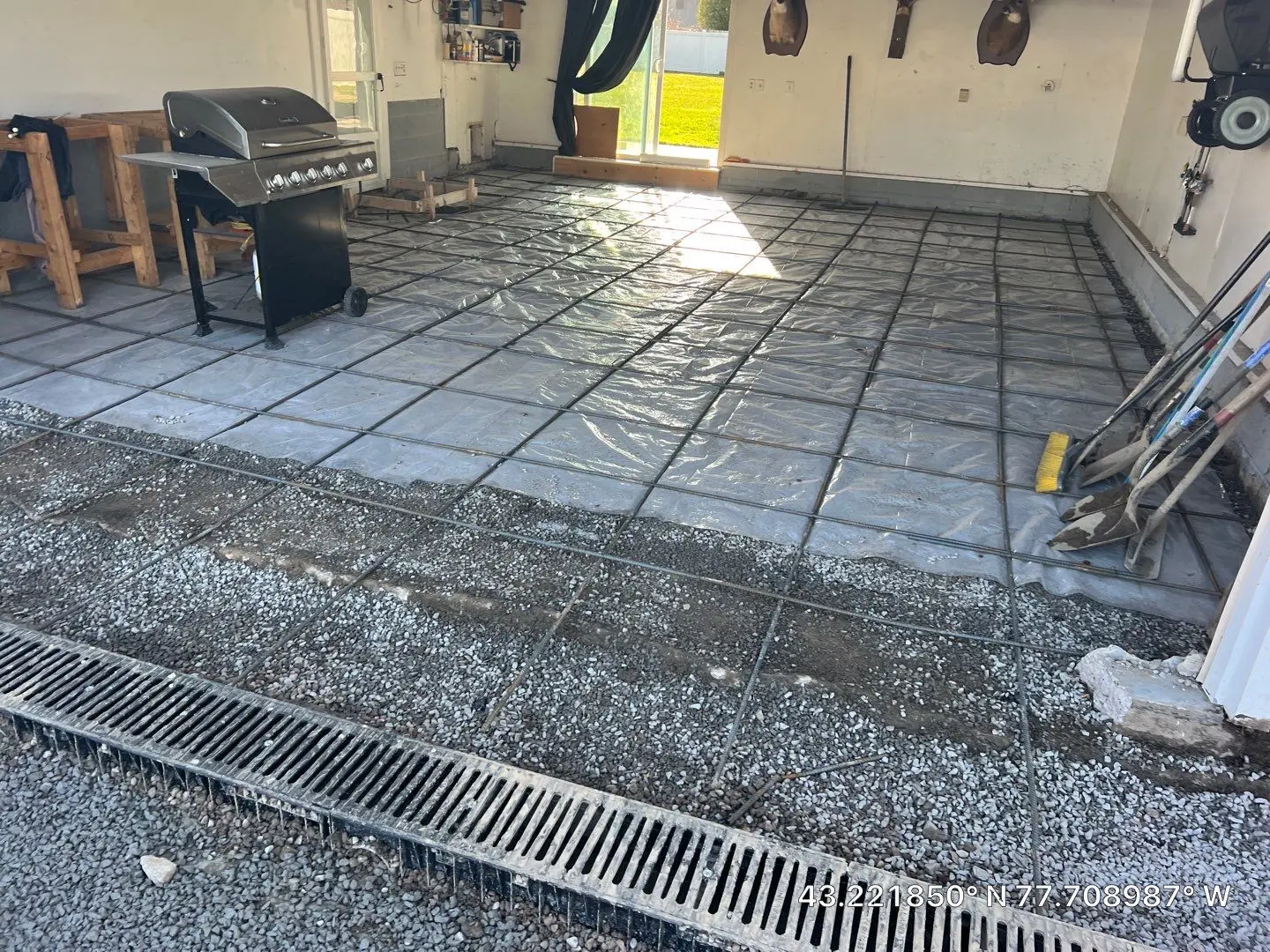 Garage floor prep in Greece NY showing the compacted stone base, vapor barrier, rebar grid on chairs, and the new trench drain at the door