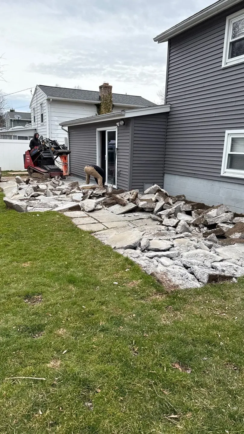 Demolition of the back patio in Greece NY, working along the side of the house toward the sliding door with the broken slab stacked for removal