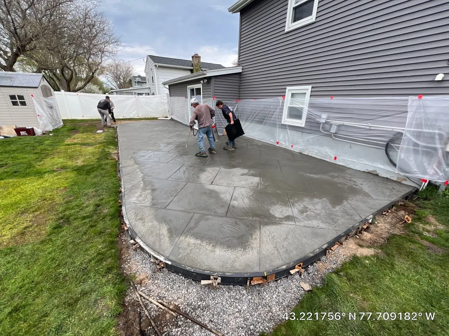 Crew applying release across the freshly stamped concrete patio in Greece NY with the siding and fence masked in plastic